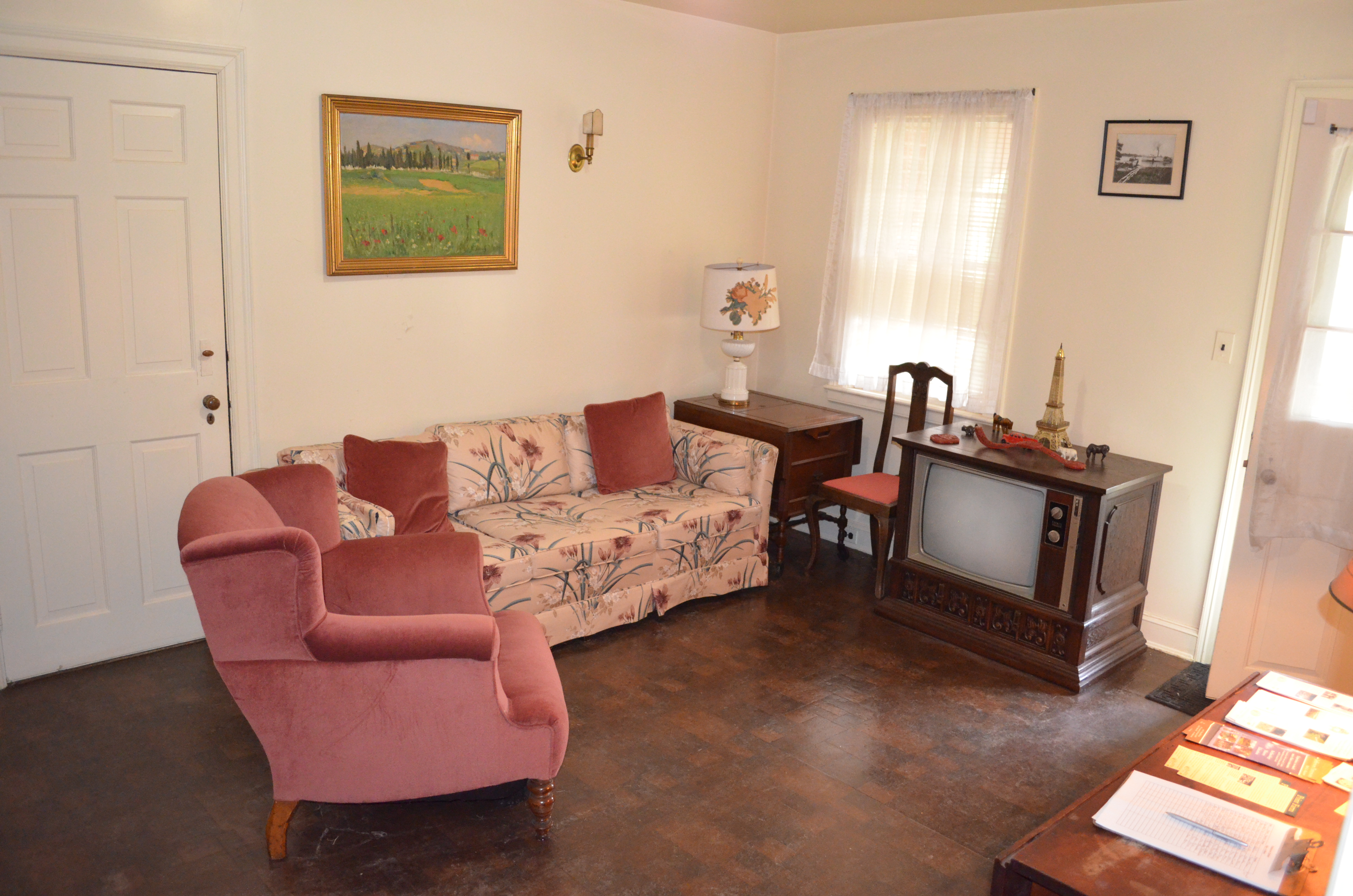 Photograph of interior room, with television unit on floor, sofa with floral pattern, and pink armchair. 